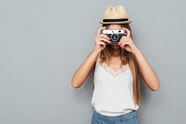 Jonge blonde vrouw die in hoed foto met camera maakt die op een grijze achtergrond wordt geïsoleerd