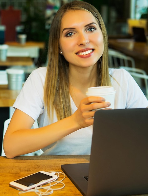 Jonge blonde vrouw die koffiethee drinkt en laptop gebruikt in een coffeeshop