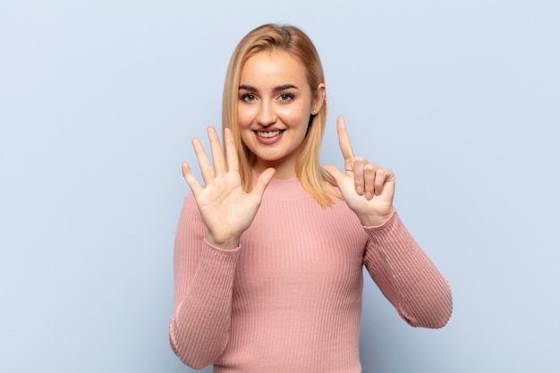 Jonge blonde vrouw die lacht en er vriendelijk uitziet, nummer zeven of zevende toont met de hand naar voren, aftellend