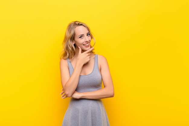 Jonge blonde vrouw die lacht, genietend van het leven, zich gelukkig, vriendelijk, tevreden en onbezorgd voelt met de hand op de kin tegen de oranje muur