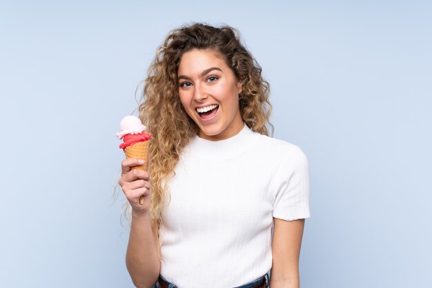 Jonge blonde vrouw die met krullend haar een cornetroomijs houdt dat op blauw met verbazing en geschokte gezichtsuitdrukking wordt geïsoleerd