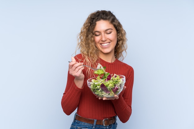 Jonge blonde vrouw die met krullend haar een salade over muur houdt