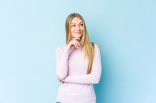 Jonge blonde vrouw die op blauwe achtergrond wordt geïsoleerd die zijwaarts met twijfelachtige en sceptische uitdrukking kijkt.