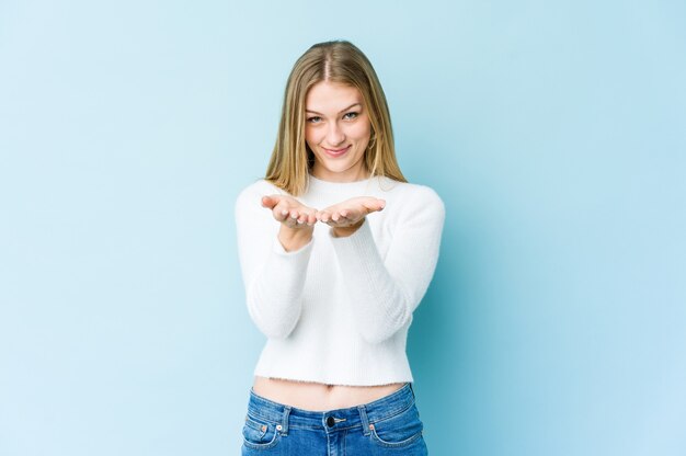 Jonge blonde vrouw die op blauwe muur wordt geïsoleerd die iets met palmen houdt, aan camera aanbiedt