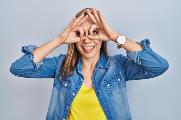 Jonge blonde vrouw die over een blauwe achtergrond staat en een goed gebaar doet, zoals een verrekijker die de tong uit de ogen steekt en door de gekke uitdrukking van de vingers kijkt