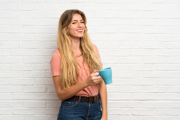 Jonge blonde vrouw die over witte bakstenen muur hete kop van koffie houdt