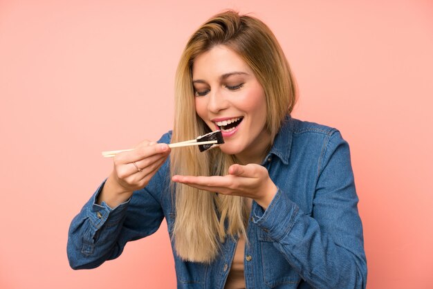 Jonge blonde vrouw die sushi over roze muur eet
