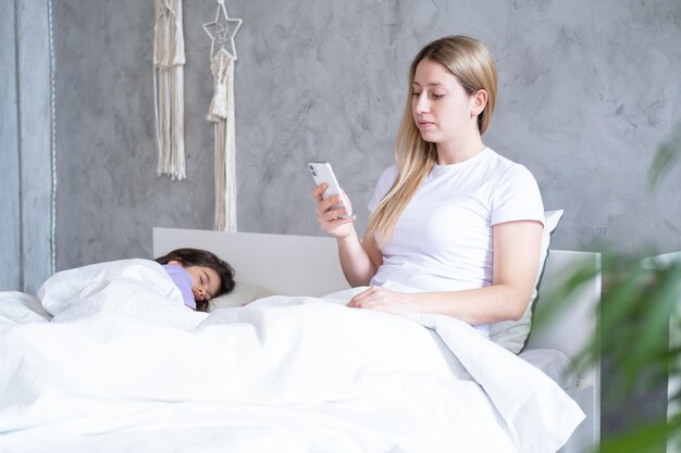 Jonge blonde vrouw die telefoon gebruikt die op bed ligt met haar slapende schattige kinddochter