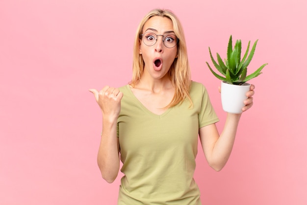 Jonge blonde vrouw die verbaasd kijkt in ongeloof en een cactus vasthoudt