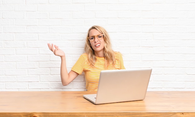 Jonge blonde vrouw die verbaasd, verward en gestrest kijkt, zich tussen verschillende opties afvraagt, zich onzeker voelt met behulp van een laptop