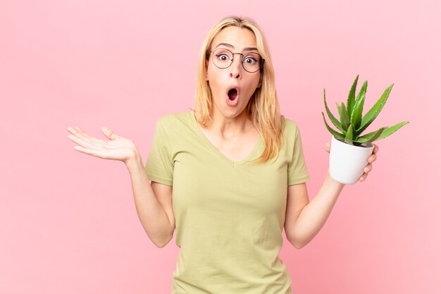 Jonge blonde vrouw die verrast en geschokt kijkt, met open mond terwijl ze een object vasthoudt en een cactus vasthoudt