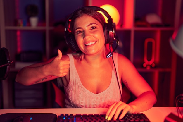 Jonge blonde vrouw die videogames speelt met een koptelefoon en een gelukkig duim omhoog gebaar met de hand doet. goedkeurende uitdrukking kijkend naar de camera die succes toont.