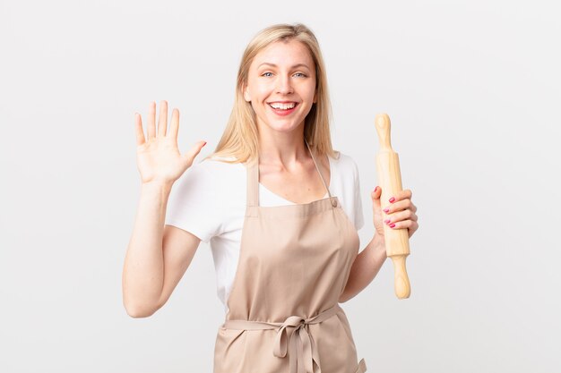 Jonge blonde vrouw die vrolijk lacht, met de hand zwaait, je verwelkomt en begroet. bakker concept