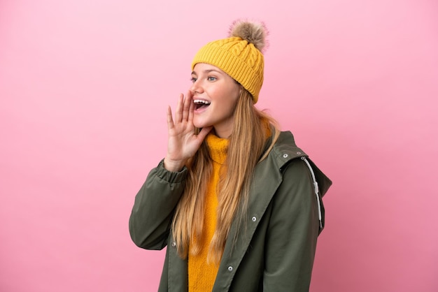 Foto jonge blonde vrouw die winterjas draagt die op roze achtergrond wordt geïsoleerd en met wijd open mond naar lateraal schreeuwt