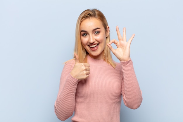 Jonge blonde vrouw die zich blij, verbaasd, tevreden en verrast voelt, oké en duimen omhoog gebaren, glimlachend