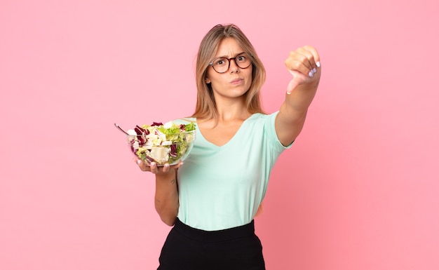 Jonge blonde vrouw die zich boos voelt, duimen naar beneden laat zien en een salade vasthoudt