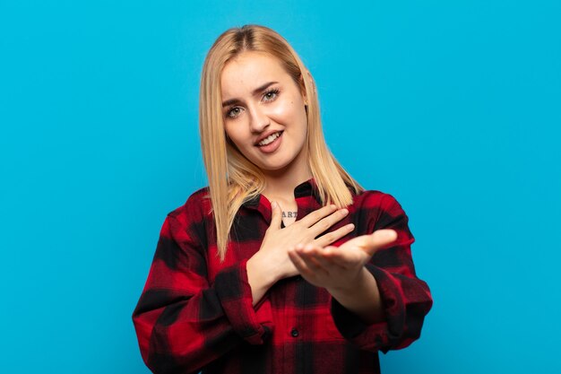 Jonge blonde vrouw die zich gelukkig en verliefd voelt, glimlachend met de ene hand naast het hart en de andere vooraan gestrekt