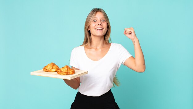 Jonge blonde vrouw die zich geschokt voelt, lacht en succes viert en een croissantblad vasthoudt