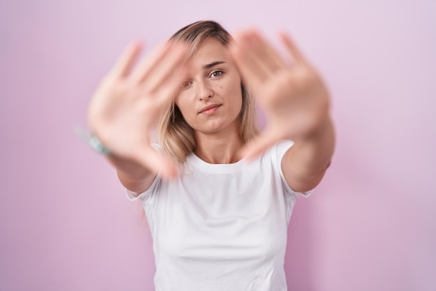 Jonge blonde vrouw die zich over roze achtergrond bevindt die frame doet met handpalmen en vingers, cameraperspectief