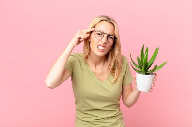 Jonge blonde vrouw die zich verward en verbaasd voelt, laat zien dat je gek bent en een cactus vasthoudt