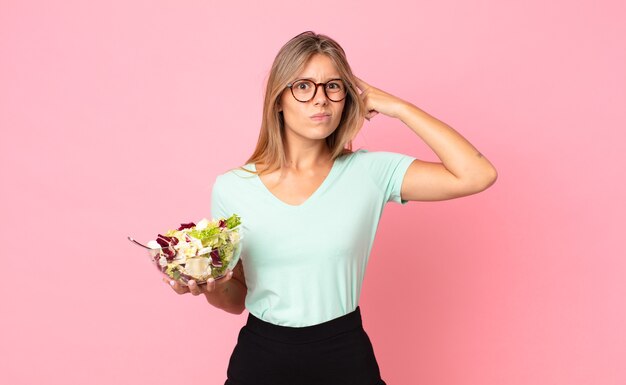 Jonge blonde vrouw die zich verward en verbaasd voelt, laat zien dat je gek bent en een salade vasthoudt