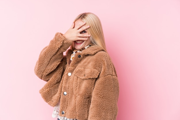 Jonge blonde vrouw draagt een jas tegen een roze muur knipperend naar de camera door vingers, beschaamd bedekkend gezicht.