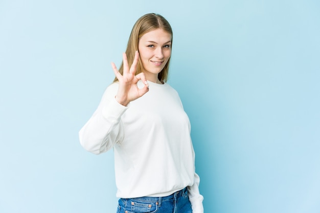 Jonge blonde vrouw geïsoleerd op blauw knipoogt en houdt een goed gebaar met de hand.