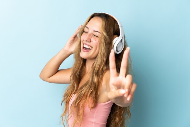 Jonge blonde vrouw geïsoleerd op blauw muziek luisteren en zingen