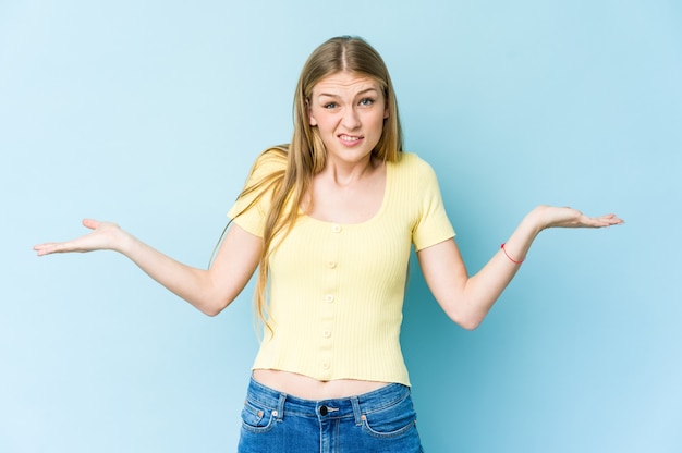 Jonge blonde vrouw geïsoleerd op blauw, verward en twijfelachtig schouders ophalen om een kopie ruimte vast te houden.