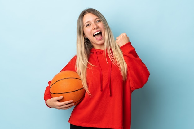 Jonge blonde vrouw geïsoleerd op blauwe muur basketbal spelen