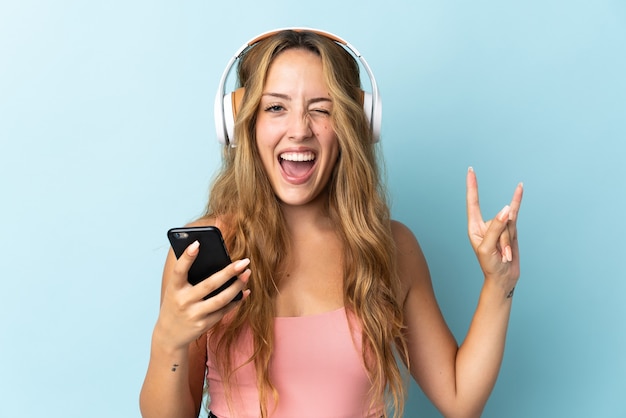 Jonge blonde vrouw geïsoleerd op blauwe muur luisteren muziek met een mobiele rock gebaar maken