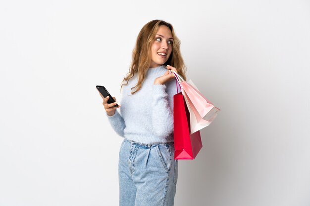 Jonge blonde vrouw geïsoleerd op een witte muur met boodschappentassen en een mobiele telefoon