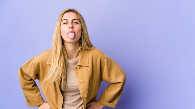 Jonge blonde vrouw geïsoleerd op paars grappig en vriendelijk tong uitsteekt.