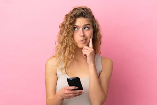 Jonge blonde vrouw geïsoleerd op roze achtergrond met behulp van mobiele telefoon en denken