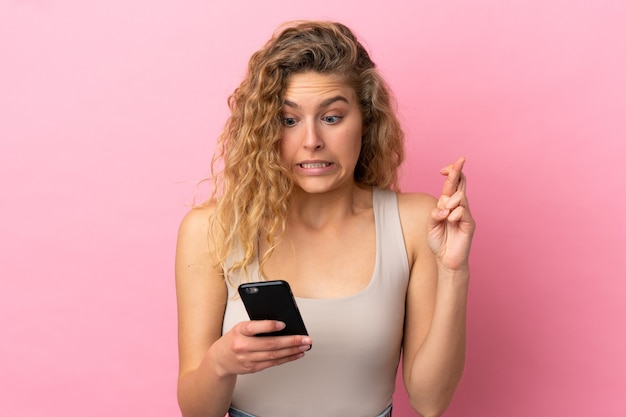 Jonge blonde vrouw geïsoleerd op roze achtergrond met behulp van mobiele telefoon met vingers over elkaar