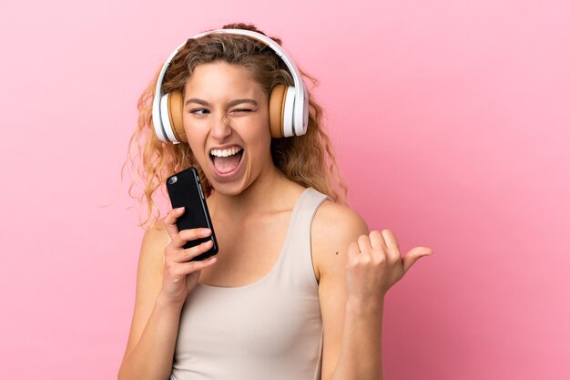 Jonge blonde vrouw geïsoleerd op roze achtergrond muziek luisteren met een mobiel en zingen