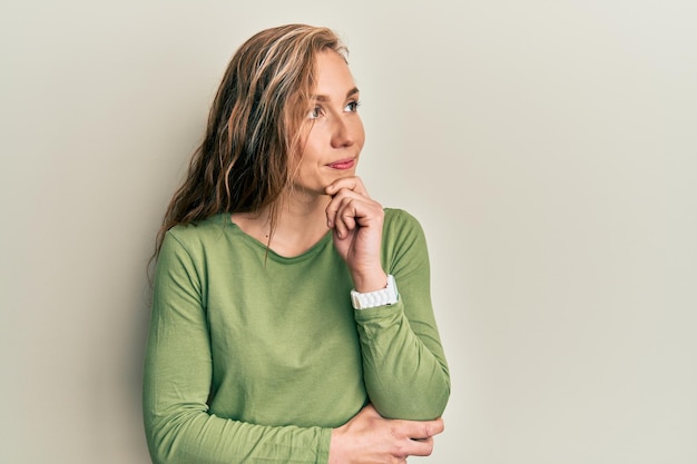 Jonge blonde vrouw, gekleed in casual kleding, denkend geconcentreerd over twijfel met de vinger op de kin en opkijkend, zich afvragend