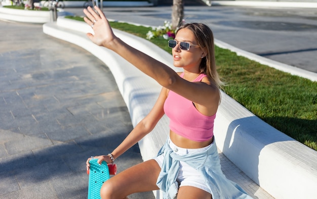 Jonge blonde vrouw gekleed in stijlvolle kleding en zonnebril met skateboard buiten op zonnige zomerdag