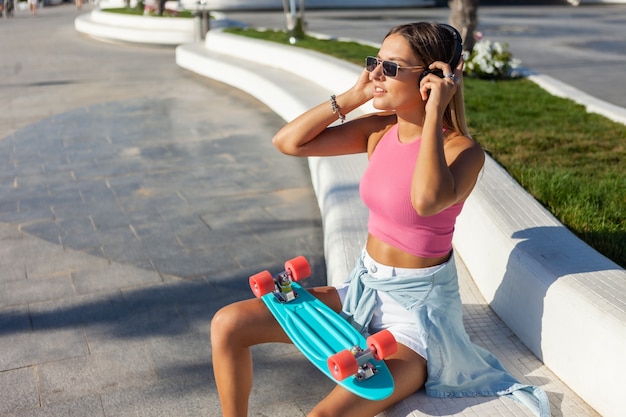 Jonge blonde vrouw gekleed in stijlvolle kleding en zonnebril met skateboard en koptelefoon buiten op zonnige zomerdag
