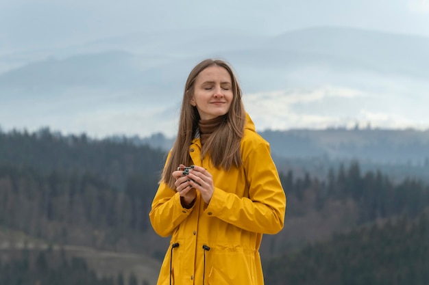 Jonge blonde vrouw genieten van het berglandschap Meisje in gele jas met kopje thee op de bergen achtergrond