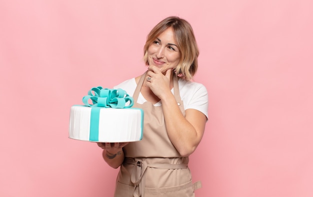 Jonge blonde vrouw glimlachend met een gelukkige, zelfverzekerde uitdrukking met de hand op de kin, zich afvragend en opzij kijkend