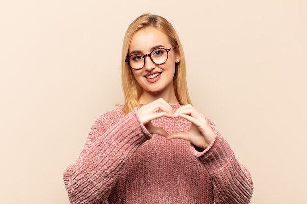 Jonge blonde vrouw glimlacht en voelt zich gelukkig, schattig, romantisch en verliefd, hartvormend met beide handen