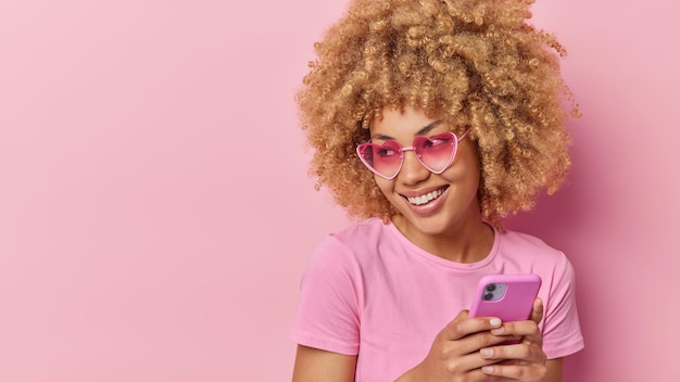 Jonge blonde vrouw glimlacht gelukkig kijkt weg stuurt bericht via smartphone draagt casual t-shirt en hartvormige zonnebril poses tegen roze achtergrond met lege ruimte voor uw reclame