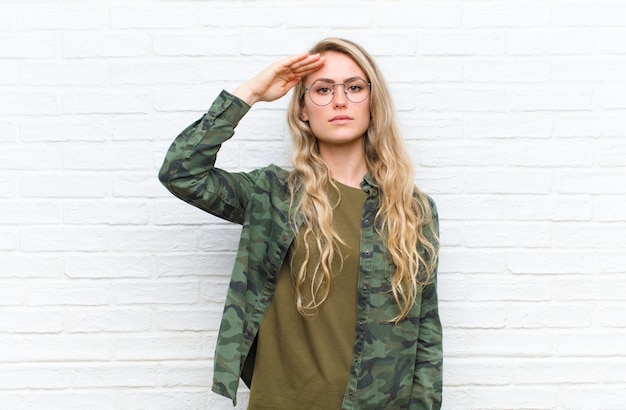 Jonge blonde vrouw groet met een militaire groet in een daad van eer en patriottisme, respect tonen tegen bakstenen muur