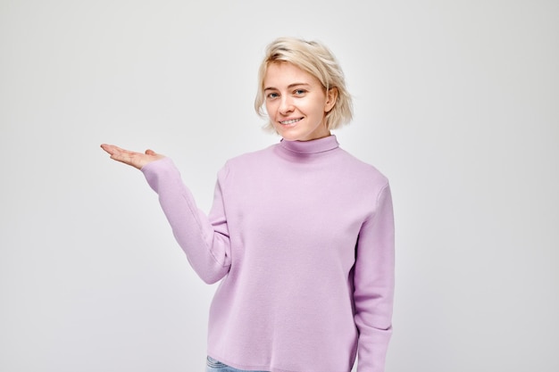Jonge blonde vrouw in casual met iets in de hand die lege ruimte voor product of tekst demonstreert geïsoleerd op witte studio achtergrond