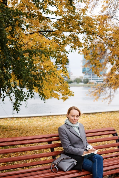 Jonge blonde vrouw in elegante kleding zittend onder herfstgebladerte in stadspark