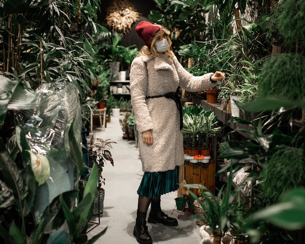 Foto jonge blonde vrouw in gebreide muts en namaakbontjas kiest vaste plant in bloemenwinkel, bloem in pot, nieuw normaal concept, met beschermend medisch masker, blanke 30 jaar oud