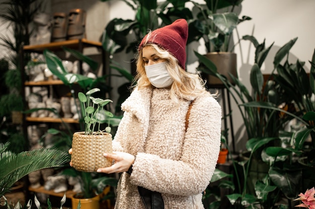 Jonge blonde vrouw in namaakbontjas kiest bloem in rieten pot bij bloemenwinkel. In de hand houden, gebreide muts en beschermend medisch masker dragen, nieuwe normen van het sociale leven, hobby's