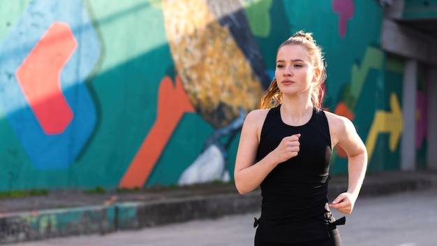 Jonge blonde vrouw in sportkleding draait op de weg bij buiten training, veelkleurige muur op de achtergrond