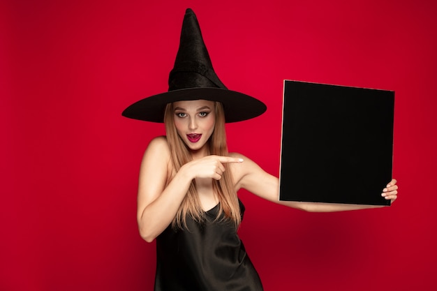 Jonge blonde vrouw in zwarte hoed en kostuum op rode achtergrond. aantrekkelijke kaukasische vrouwelijke model poseren. halloween, zwarte vrijdag, cybermaandag, verkoop, herfstconcept. kopieerruimte. houdt zwarte plaat vast.